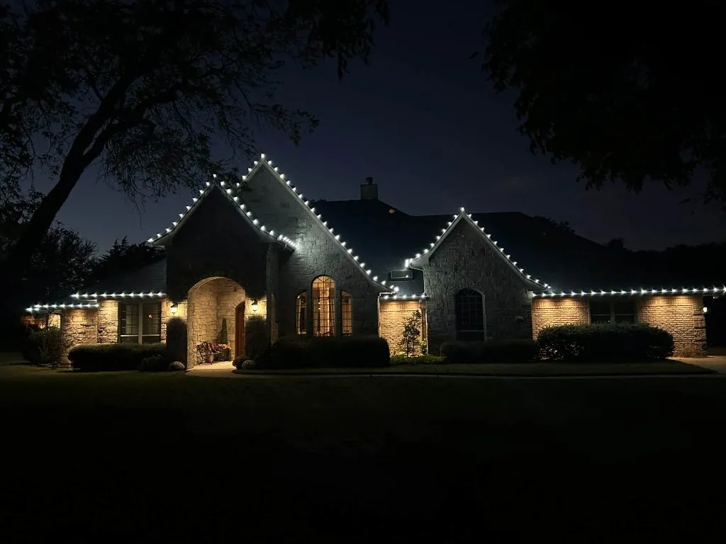 Holiday lighting installed along the roofline of a stone home at night